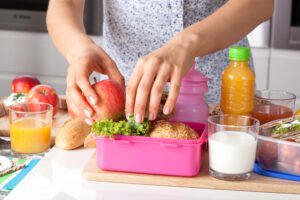 A person is packing a pink lunchbox with an apple, lettuce, and bread, surrounded by drinks and snacks on a kitchen counter.
