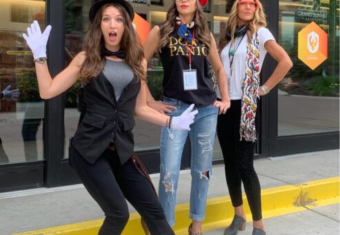 Three women dressed in creative costumes, including hats and gloves, posing energetically outside a venue during a Corporate Film Festival team building event, showcasing their excitement and team spirit.