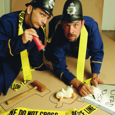 Two actors dressed as police officers humorously investigate a mock crime scene in a playful setup for a Murder Mystery Dinner event. They are kneeling on the ground, surrounded by yellow police line tape, with a chalk outline on the floor resembling the shape of a duck. One officer is holding a red flashlight while the other points at a toy duck with a sign labeled 'Dead Duck.' The scene is lighthearted and comedic, emphasizing the fun, interactive nature of the mystery event. thumbnail