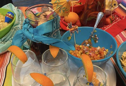 A festive spread featuring glasses garnished with orange slices, a pitcher of colorful drink, fresh salsa, and bowls of chips, all part of the Salsa Showdown team building event.