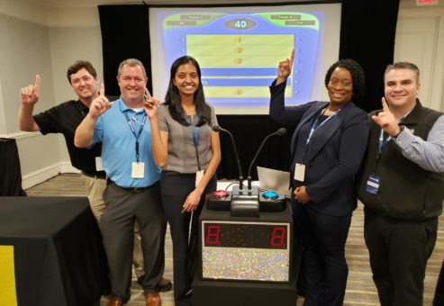 A team of five participants posing with enthusiasm and pointing to the screen, showing a game show setup during the Corporate Feud team building activity, highlighting team spirit, competition, and fun.