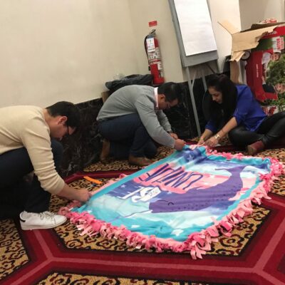 Team members collaborate during the Hope for the Holidays event, carefully crafting a fleece blanket to donate to local seniors as part of their charitable efforts. thumbnail
