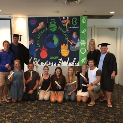 A group of participants proudly posing in front of their completed mural, created during The Big Picture team building event. The mural features words like 