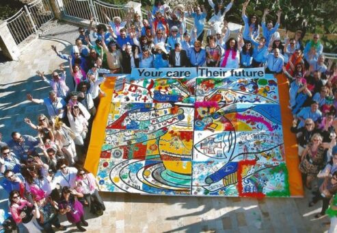 A large group of participants gathers around a completed mural during The Big Picture team building event, celebrating their collaborative artwork with hands raised in excitement. The vibrant mural emphasizes teamwork and shared goals.