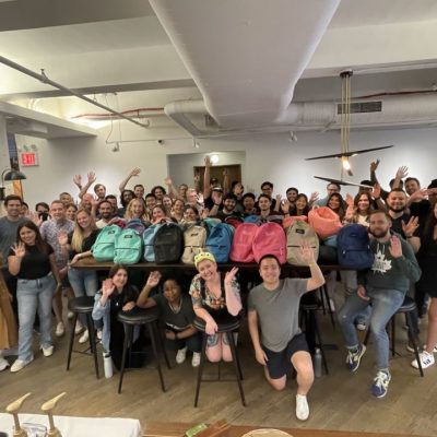 Large group of smiling participants posing with colorful backpacks filled with school supplies after a successful corporate csr event, highlighting teamwork and community impact through the Tools for Schools program. thumbnail