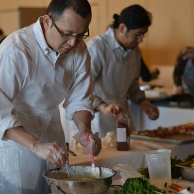A participant at the 'Food Truck Challenge' is preparing a dish. Wearing an apron and gloves, he focuses on mixing ingredients in a large bowl. Behind him, other participants are also engaged in their culinary tasks, surrounded by fresh ingredients thumbnail