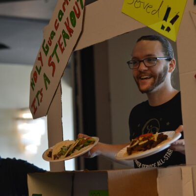 Participants enthusiastically showcase their creative food truck setup during a 'Food Truck Challenge' team building event. A team member stands under a handcrafted food truck façade, proudly presenting plates of tacos. thumbnail