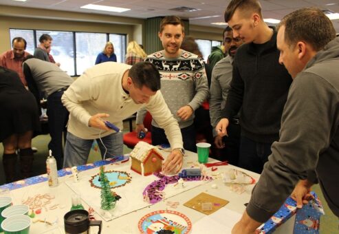 A group of men working together on decorating a gingerbread house during a team building event. One man uses a hot glue gun to assemble pieces, while others discuss and add decorations to the house on a festive, holiday-themed table.