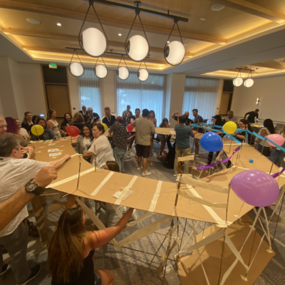 Teams collaborate to construct a large bridge decorated with balloons during the Bridging the Divide team building activity. thumbnail