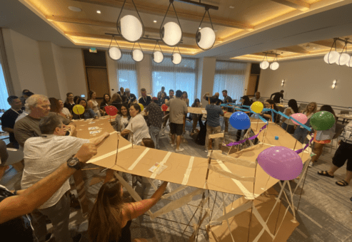 Teams collaborate to construct a large bridge decorated with balloons during the Bridging the Divide team building activity.