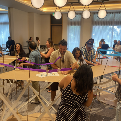 Teams work together to build a large bridge structure using cardboard and colorful materials during the Bridging the Divide team building activity. thumbnail