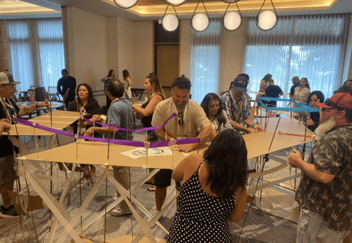 Teams work together to build a large bridge structure using cardboard and colorful materials during the Bridging the Divide team building activity.