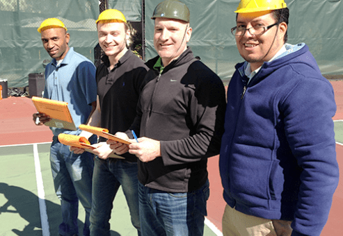 Four team members wearing yellow safety helmets, standing together outdoors on a tennis court, smiling and holding team building materials during the 'Catapult to Success' team building activity.