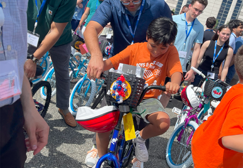 A group of volunteers assisting children in riding their newly assembled bikes during a Charity Bike Build event. A boy in an orange shirt excitedly rides a decorated blue bike, guided by a volunteer.
