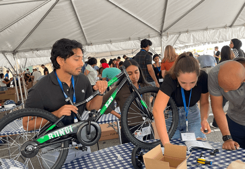 A group of participants working together to assemble a green bicycle during a Charity Bike Build event. They are under a large tent, focused on attaching parts with tools. Other groups in the background are also working on assembling bikes.