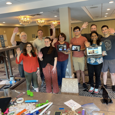 Group of participants proudly waving after completing their Domino Effect team building activity, surrounded by creative contraption materials. thumbnail