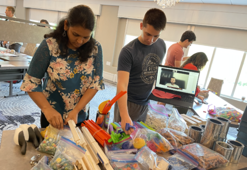 Participants sorting through materials and building supplies during the Domino Effect team building event, with a remote team member visible on a tablet.