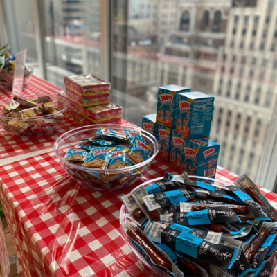 Snacks and food items, including Skippy peanut butter packs and granola bars, arranged on a red checkered table at a corporate charitable team building event. thumbnail