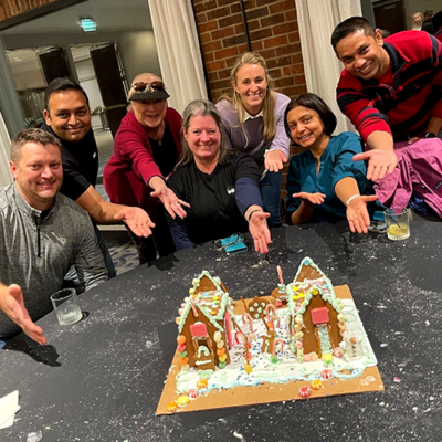 A group of seven enthusiastic participants proudly presenting their gingerbread house creation during a team building event. The gingerbread structure is decorated with icing, candy canes, and colorful candies. The team members are smiling and extending their hands toward the gingerbread house. thumbnail