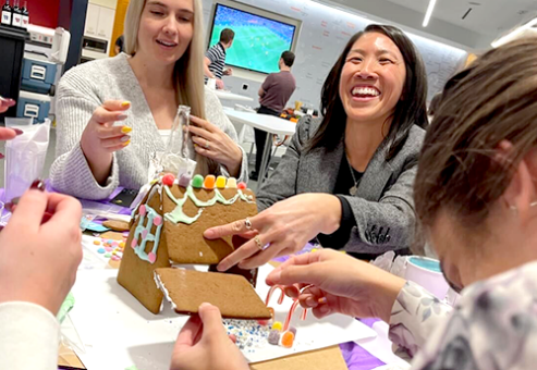 Two women are laughing and enjoying a gingerbread house competition during a team building event. They are actively working together to construct and decorate a small gingerbread house with colorful candies. The setting appears to be a bright, modern office space, and other team members can be seen in the foreground, contributing to the creative project.