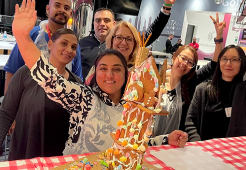 A group of six people enthusiastically posing next to their gingerbread house creation during a team building competition. The gingerbread structure is tall and decorated with colorful candies, icing, and festive embellishments.