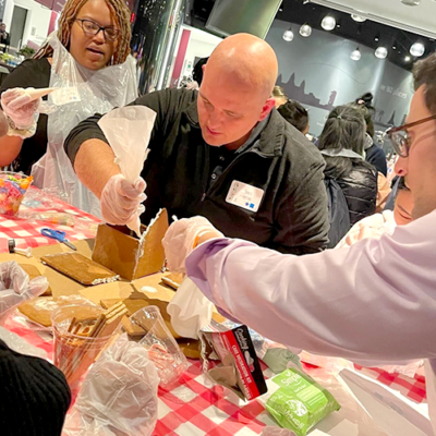 A team collaborating on constructing a gingerbread house during a team building event. One participant is applying icing with a piping bag to secure the house's walls, while others assist by holding the pieces together. The table is covered with a red and white checkered cloth, and various gingerbread house components and decorations are visible. The atmosphere is lively, with more participants in the background working on similar projects. thumbnail