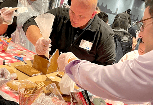 A team collaborating on constructing a gingerbread house during a team building event. One participant is applying icing with a piping bag to secure the house's walls, while others assist by holding the pieces together. The table is covered with a red and white checkered cloth, and various gingerbread house components and decorations are visible. The atmosphere is lively, with more participants in the background working on similar projects.