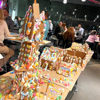 A display of intricately decorated gingerbread houses lined up during a team building competition. The houses are adorned with colorful candies, icing, and sprinkles, with one particularly tall gingerbread structure standing out in the foreground. thumbnail