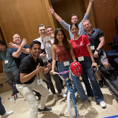 A group of smiling participants posing proudly with a completed bicycle during a Charity Bike Build event. Some team members are giving thumbs up and raising their hands in excitement, celebrating their achievement after assembling the bike. thumbnail