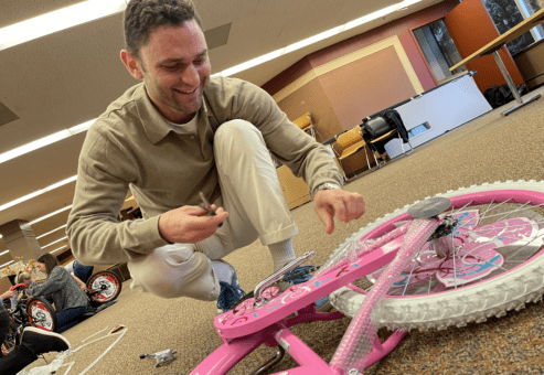 A man crouches on the floor with a smile, carefully assembling a pink bicycle during a Charity Bike Build team building event. He uses tools to attach the final pieces, showcasing hands-on involvement in creating bikes for children.
