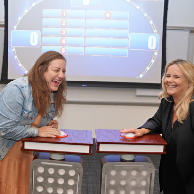 Two participants laughing and enjoying themselves while standing at game show podiums with buzzers, ready to answer a question during the Corporate Feud team building event, with a game board visible in the background. thumbnail
