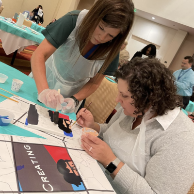 Two participants collaborate on painting a section of a mural during The Big Picture team building event. Wearing aprons and gloves, they focus on adding fine details to their canvas, contributing to the larger masterpiece. thumbnail
