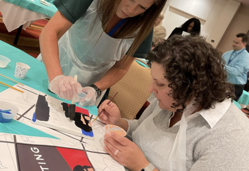 Two participants collaborate on painting a section of a mural during The Big Picture team building event. Wearing aprons and gloves, they focus on adding fine details to their canvas, contributing to the larger masterpiece.